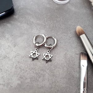 Può includere: Un paio di orecchini a cerchio d'argento con un ciondolo a forma di stella di David. Gli orecchini sono su una superficie grigia.