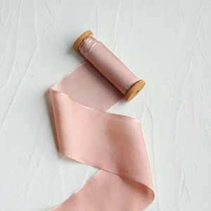 May include: A spool of blush pink silk ribbon with a wooden core. The ribbon is unraveled, showing its soft texture and colour. The wooden spool is a light brown colour. The ribbon is suitable for crafts, gift wrapping, or decorative purposes.