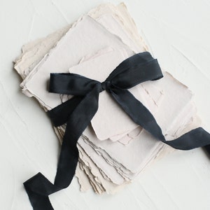 May include: A stack of handmade, textured paper in shades of pink and beige, tied together with a black satin ribbon.