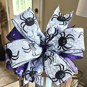 Pode incluir: Decoração com tema de Halloween com uma lanterna de madeira encimada por um grande laço. O laço tem fita branca com desenhos de aranhas e teias de aranha pretas e detalhes em fita roxa. A lanterna tem uma janela de vidro.