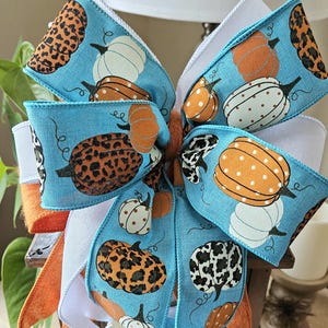 May include: A decorative bow with a light blue background and a pumpkin pattern. The pumpkins are orange, white, and leopard print. The bow is made of ribbon and has white and orange accents. Perfect for fall decor.