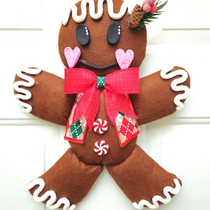 May include: A brown felt gingerbread man Christmas decoration with white icing details. Features black eyes, pink glitter hearts, a red bow, and candy buttons. Adorned with a pine cone and berries. Festive holiday decor.