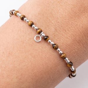 May include: A beaded bracelet featuring alternating brown tiger eye gemstone beads and small polished silver-tone metal spheres. The bracelet includes a single small silver-tone metal ring charm suspended from the center of the beaded strand.