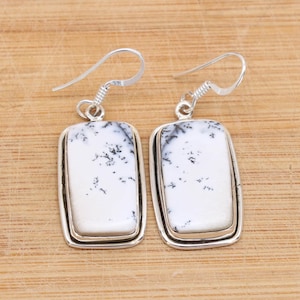 May include: A pair of rectangular dangle earrings. Each earring features a white stone with black speckles, set in a silver-colored frame. The earrings have a simple hook design for pierced ears. The background is a light brown wooden surface.