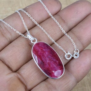 May include: A silver necklace featuring a large, oval-shaped, faceted ruby pendant. The ruby is a deep, vibrant red color and is set in a silver bezel. The chain is a delicate silver link chain with a lobster clasp.