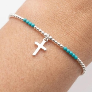 Peut inclure: Un bracelet élastique délicat composé de petites perles argentées polies alternant avec une section de perles bleu turquoise vif. Une breloque en forme de croix argentée, simple et lisse, est suspendue au centre du bracelet.