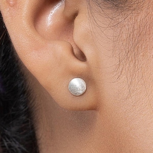 May include: A close-up of a person's ear with a small, round, silver stud earring. The earring has a textured surface and is set against the skin. The person's dark hair is visible in the background.