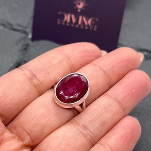 May include: A silver ring featuring a large oval ruby gemstone. The ruby is a deep red colour.