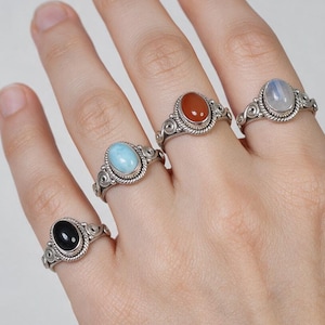 May include: Four silver rings featuring oval-shaped cabochon gemstones in black, light blue, reddish-orange, and iridescent white. Each ring has a twisted rope border around the stone and decorative scrollwork details on the band.