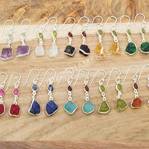 Op de afbeelding: Een verzameling zilveren oorbellen met diverse kleurrijke edelsteenhangers. De oorbellen hebben een haakontwerp en zijn versierd met amethist, kwarts, granaat en andere stenen. De oorbellen worden gepresenteerd op een houten oppervlak.