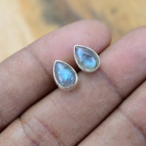 May include: A pair of teardrop-shaped stud earrings. Each earring features a blue and grey iridescent stone set in a silver bezel. The earrings are displayed on a light brown surface.