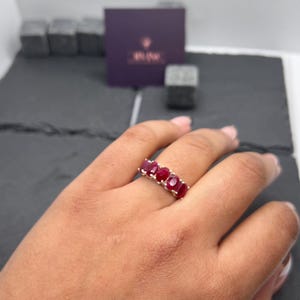 Può includere: Un anello dorato con cinque pietre preziose rosse ovali. L'anello è indossato su una mano, evidenziando il colore delle pietre e l'elegante design. Sullo sfondo, una scatola viola e una superficie scura.