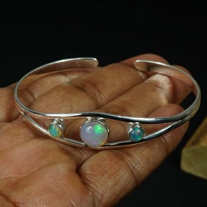 May include: A silver cuff bracelet featuring three opal gemstones. The bracelet has a simple, elegant design with a polished finish. The opals display a range of colors, including green, blue, and white, adding a touch of sparkle.