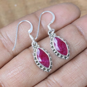 May include: A pair of silver drop earrings. Each earring features a marquise-cut, deep red gemstone set within a beaded silver frame. The earrings have a simple hook closure. The background is neutral.