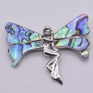 May include: A silver-toned fairy brooch with iridescent abalone wings. The fairy figure is in a seated position with flowing lines. The wings display shades of blue, green, and purple. This jewelry piece is a unique accessory.