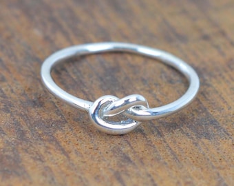 May include: A silver knot ring with a simple design. The ring is made of a thin band of metal and features a knot in the center.