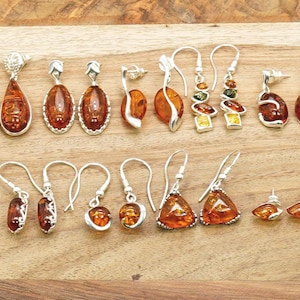 May include: A selection of amber earrings with silver settings. The earrings showcase diverse shapes, such as teardrops, ovals, and triangles. The amber stones vary in colour from light to dark brown, displayed on a wooden surface.