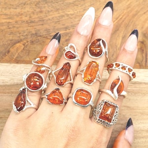Peut inclure: Plusieurs bagues en argent ornées de pierres d'ambre de différentes formes. Les bagues présentent divers designs, notamment des montures en ambre ovales, rectangulaires et en forme de goutte. Les pierres d'ambre varient du brun clair au brun foncé.