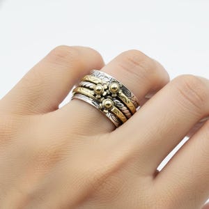 Könnte beinhalten: Silber- und goldfarbener Spinner-Ring mit floralem Design. Der Ring hat mehrere Bänder, mit goldfarbenen und silberfarbenen Bändern. Der Ring hat ein florales Design mit kleinen goldfarbenen Blumenakzenten. Der Ring befindet sich an einem Finger.