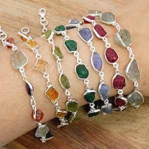 Op de afbeelding: Verschillende zilveren kettingarmbanden met kleurrijke, onregelmatig gevormde edelstenen. De stenen hebben verschillende kleuren, waaronder groen, oranje, paars en rood. De armbanden zijn met elkaar verbonden en lijken om een pols te worden gedragen.