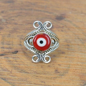 May include: A silver ring with a red and white evil eye centrepiece. The ring has detailed silverwork around the eye, including swirls and beaded patterns. The eye itself is a bright red with a white and black pupil.