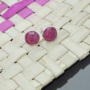 May include: A pair of silver stud earrings with pink gemstone settings. The earrings are on a woven background.