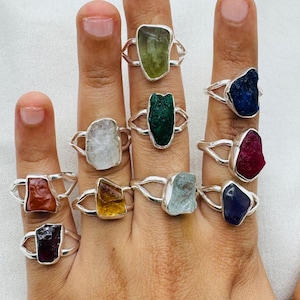 Op de afbeelding: Een set van tien zilveren ringen met verschillende gekleurde edelstenen. De edelstenen zijn ruw en onbewerkt en zijn gezet in een eenvoudige zilveren band. De ringen zijn allemaal van verschillende grootte en zijn gerangschikt in een rij op een hand.