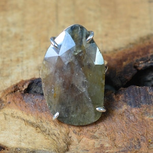 May include: A faceted, teardrop-shaped gemstone ring. The large, light-brown stone is held in place by four silver prongs. The ring is set against a natural wood background.