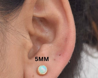 May include: A pair of opal stud earrings with a gold setting. The earrings are 5mm in diameter. The earrings are shown in a person's ear.