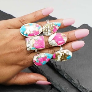 May include: A collection of five rings featuring polished stones with vibrant pink, bright blue, and white matrix patterns. The rings are set in silver-toned metal and vary in shape, including oval, rectangular, teardrop, and circular designs.