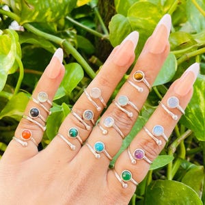May include: A hand shows a selection of silver rings, each with a different coloured gemstone. The gemstones include orange, black, green, blue, and purple. The rings are set against a background of green leaves.