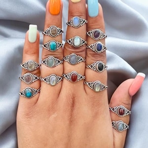 Op de afbeelding: Een hand draagt ​​een verscheidenheid aan zilveren ringen met verschillende gekleurde edelstenen. De ringen zijn allemaal in een vergelijkbare stijl, met een delicaat, versierd ontwerp. De edelstenen zijn allemaal ovaalvormig en zijn in het midden van de ringen gezet.