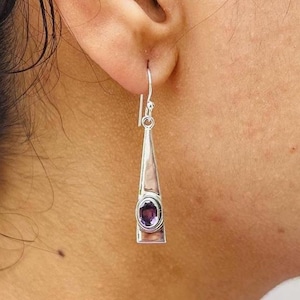 May include: A silver drop earring featuring a long, triangular metal frame with a polished finish. An oval-shaped, deep purple amethyst gemstone is bezel-set in the centre of the lower portion of the earring, which hangs from a simple silver hook.
