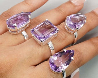 Puede incluir: Cuatro anillos de plata con grandes piedras de amatista púrpura facetadas. Las piedras preciosas tienen varias formas, incluyendo pera, rectangular y ovalada. Los anillos se muestran en una mano.