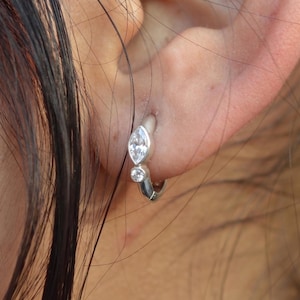 May include: A silver hoop earring featuring a marquise-cut clear gemstone and a smaller round-cut clear gemstone set along the front of the band. The earring is secured through the earlobe, showcasing a polished metallic finish and two stones.