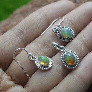 Puede incluir: Un par de pendientes de plata y un colgante con piedras preciosas de &oacute;palo de forma ovalada engastadas en un engaste de plata con un borde de cuentas. Las piedras preciosas de &oacute;palo tienen un brillo iridiscente arco&iacute;ris.
