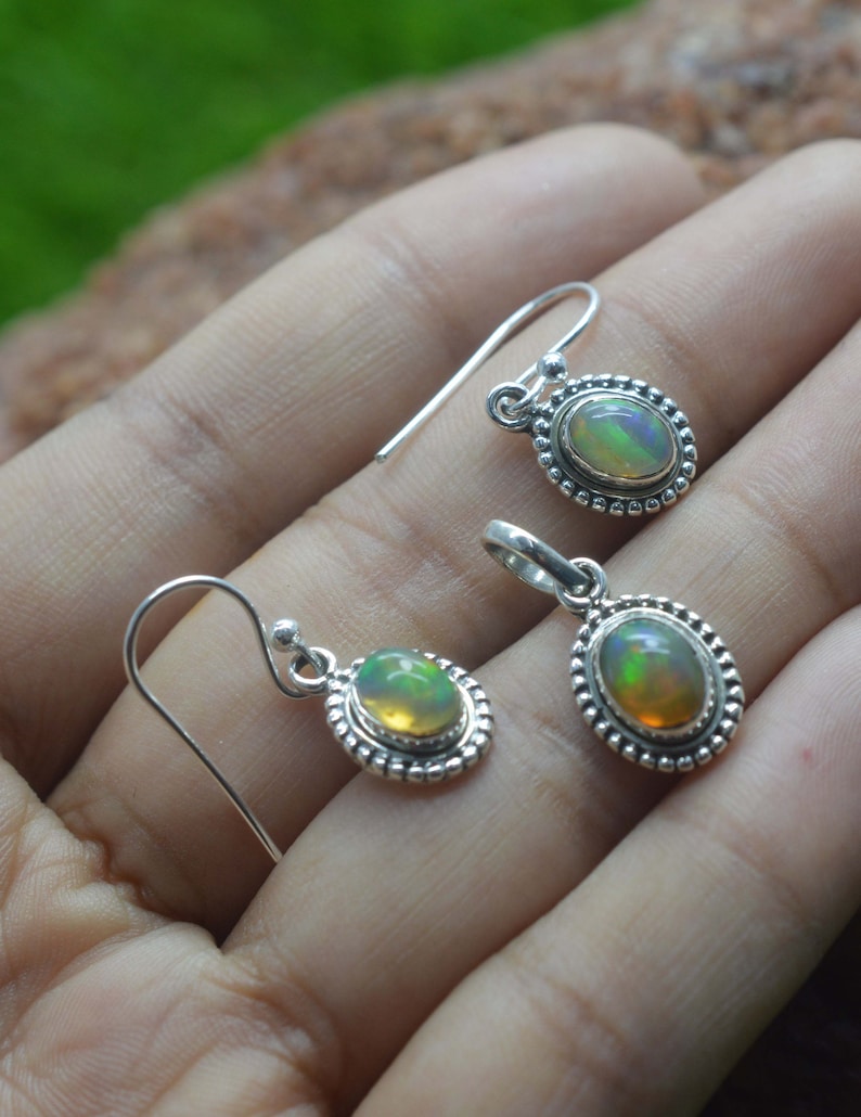 Puede incluir: Un par de pendientes de plata y un colgante con piedras preciosas de &oacute;palo de forma ovalada engastadas en un engaste de plata con un borde de cuentas. Las piedras preciosas de &oacute;palo tienen un brillo iridiscente arco&iacute;ris.