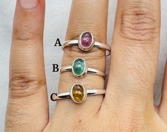 May include: Three silver rings with different colored oval gemstones. The top ring has a pink gemstone, the middle ring has a green gemstone, and the bottom ring has a yellow gemstone.