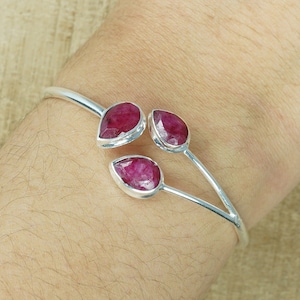 Könnte beinhalten: Ein silbernes Armreif-Armband mit drei tropfenförmigen, tiefroten Edelsteinen. Die Edelsteine sind in einzelnen Silberfassungen gefasst und über das offene Design des Armbands verteilt. Das Armband wird an einem Handgelenk getragen.