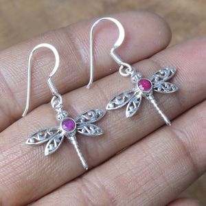 Puede incluir: Pendientes de libélula de plata con detalles de piedras preciosas rojas. Los pendientes tienen un delicado diseño de filigrana y cuelgan de un gancho.
