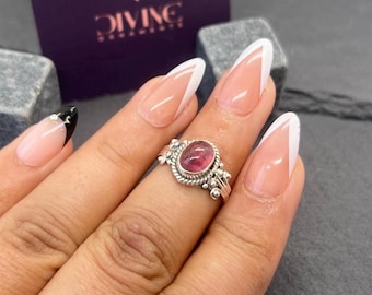 May include: A silver ring with a pink oval gemstone set in a twisted band. The ring is on a finger with a white French manicure.