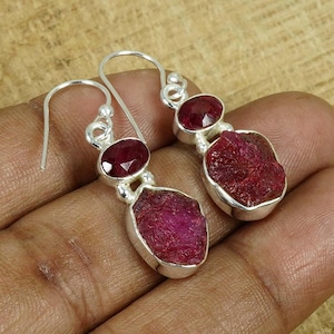 Könnte beinhalten: Ein Paar silberne Ohrringe mit zwei Rubinen. Die Ohrringe haben einen größeren, rohen Rubin und einen kleineren, facettierten Rubin.