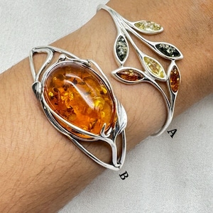 May include: Two silver bracelets featuring amber stones. The bracelet on the left has a large oval amber stone set in a silver frame with a leaf design. The bracelet on the right has a silver leaf design with multiple smaller amber stones.