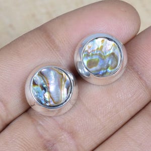Peut inclure: Une paire de boucles d'oreilles rondes avec des incrustations de coquillage d'ormeau irisé. Les boucles d'oreilles ont un sertissage en argent poli. La coquille d'ormeau affiche un mélange de couleurs bleues, vertes et dorées.