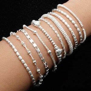 Può includere: Un assortimento di braccialetti d'argento, ognuno con un design unico, esposti su uno sfondo scuro. I braccialetti includono stili con perline, a catena e strutturati, mostrando una varietà di trame e motivi. I gioielli sono in argento lucido.