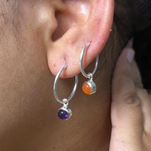 May include: A pair of silver hoop earrings, one with a purple gemstone and the other with an orange gemstone. The earrings are shown on an ear, with the gemstones dangling below the hoops. The earrings are a simple, elegant design.