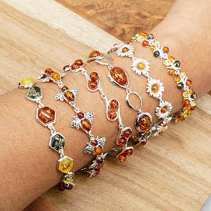 Puede incluir: Se muestran varias pulseras de plata con ámbar y elementos decorativos. Las pulseras presentan diversos diseños, incluyendo abejas, flores y formas geométricas. Las piedras de ámbar varían en color, desde el amarillo hasta el naranja oscuro, creando una estética cálida.