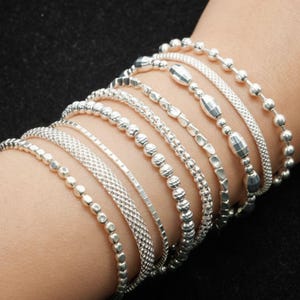 Può includere: Una collezione di braccialetti d'argento in vari design, tra cui perline, catene e stili strutturati. I braccialetti sono esposti su un polso contro uno sfondo scuro, mettendo in risalto le diverse trame e forme dei gioielli.