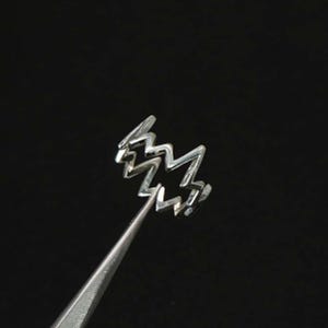 Peut inclure: Une bague argentée avec un motif en zigzag, tenue par une pince à épiler sur fond noir. Le design de la bague rappelle un éclair ou une lettre stylisée. La bague est en métal brillant.