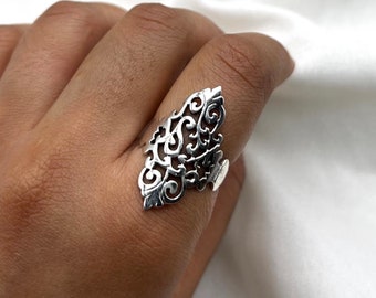 Könnte beinhalten: Ein silberfarbener Ring mit einem verzierten, durchbrochenen Design. Der Ring hat eine große, längliche Form mit komplizierten Wirbelmustern und blattartigen Details. Der Ring wird an einem Finger getragen und zeigt seine detaillierte Handwerkskunst und seinen einzigartigen Stil.